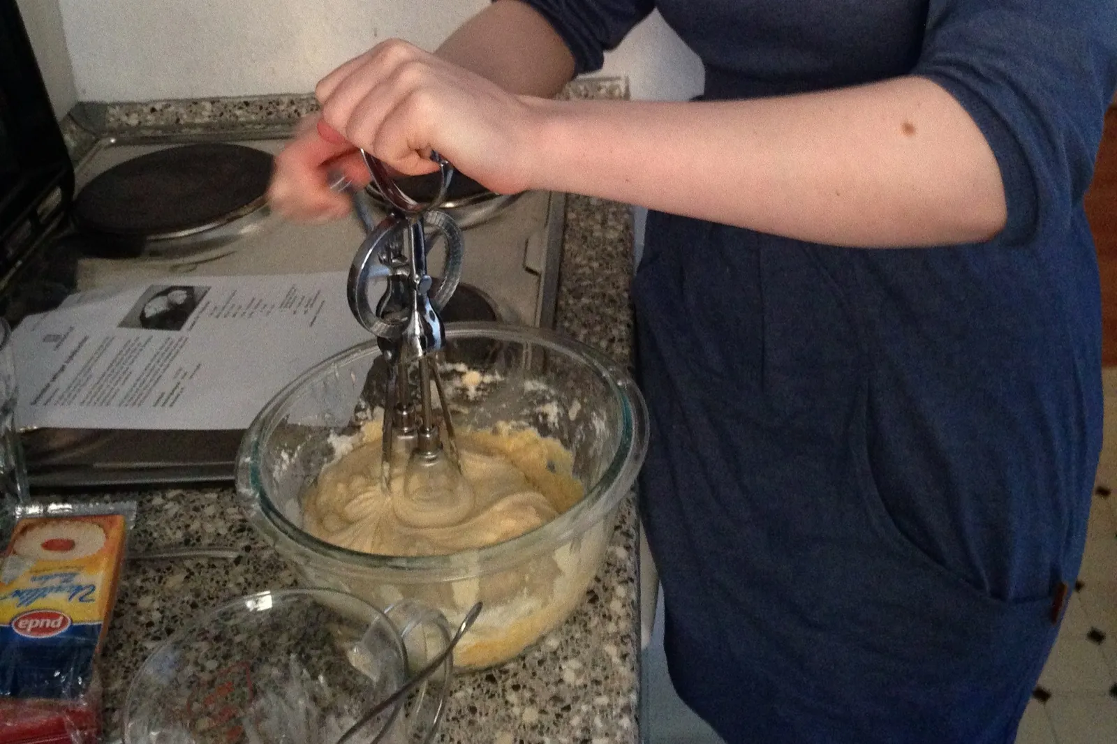 Hands working a hand-cranked mixer in a glass bowl of batter, the recipe printout beside it