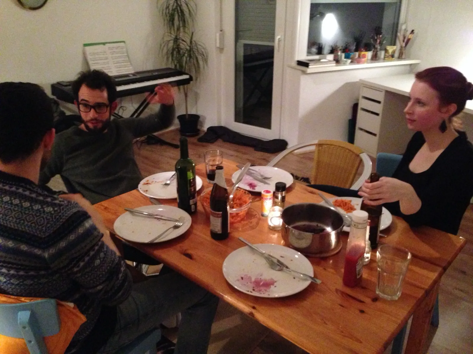Three people talking around a dinner table with empty plates, bottles and a pot