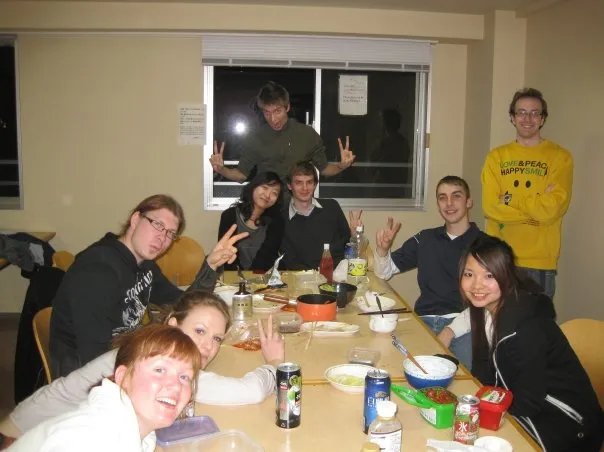 A group of students around a table covered in food and drinks in a dorm common room, several making peace signs