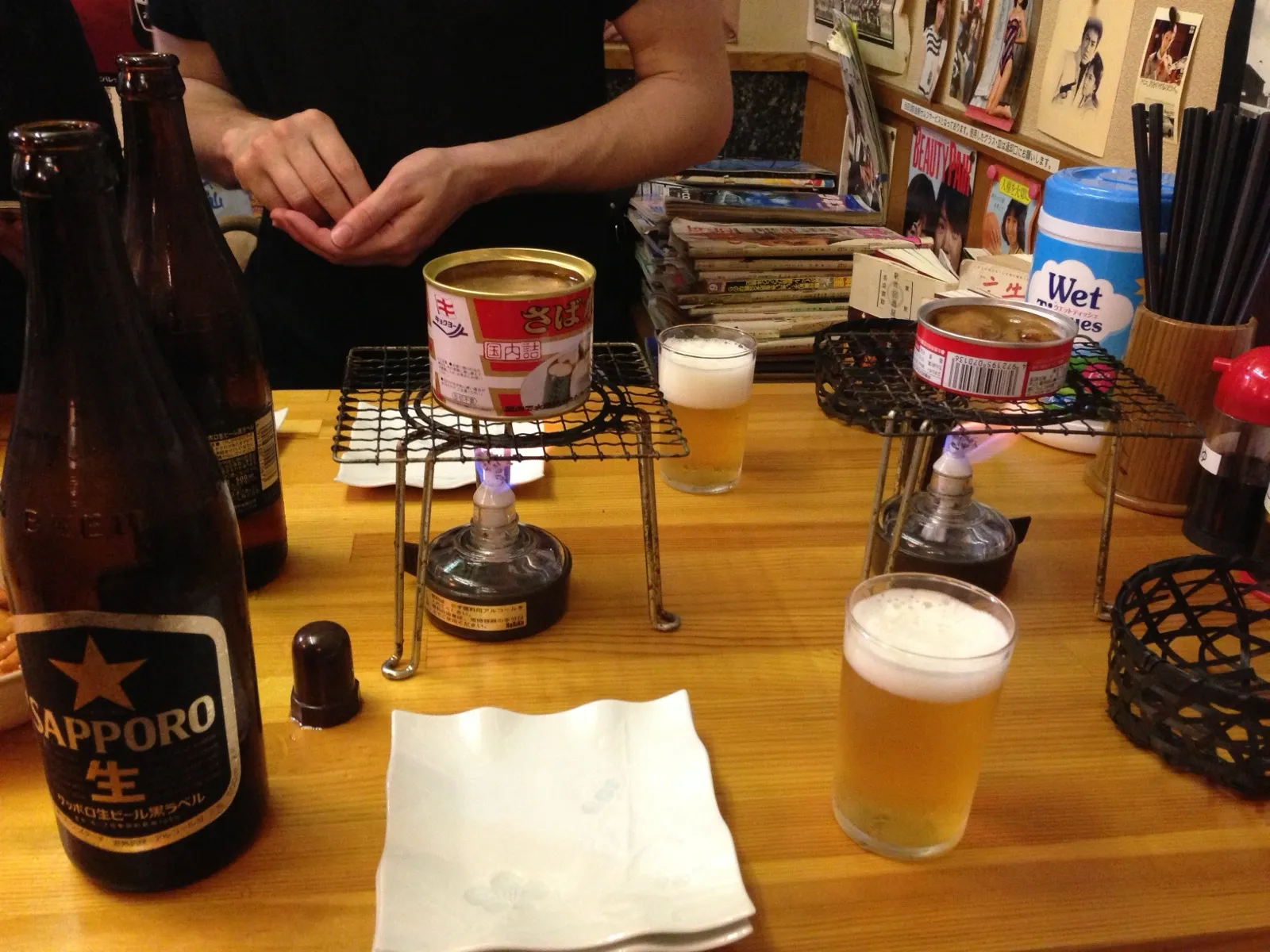 Cans of food heating on small burners on a bar counter, next to glasses of beer and a bottle of Sapporo
